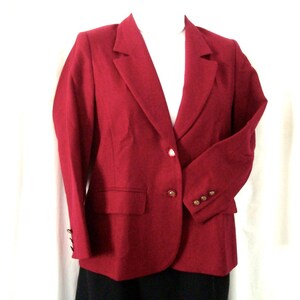 May include: A solid deep red blazer featuring a notched lapel, two front buttons, and two flap pockets. The sleeves are finished with three small gold-toned buttons at each cuff. The garment is structured with shoulder pads and a classic tailored fit.