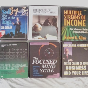 Op de afbeelding: Een verzameling van zes VHS-banden met titels over zaken en zelfverbetering. Titels zijn onder meer "Sell Your Way To The Top", "The Secrets of Power Negotiating" en "Multiple Streams of Income". De banden zitten in zwarte cassettes met kleurrijke tekst en afbeeldingen.