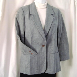 May include: A light grey blazer featuring a single-button front closure, notched lapels, and two large patch pockets on the lower front. The sleeves are finished with two small buttons at each cuff, and the garment is worn over a white turtleneck.