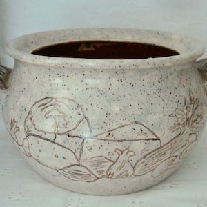 Puede incluir: Un cuenco de cerámica moteada de color blanquecino con un borde ancho y dos asas curvas. El cuenco presenta una ilustración marrón de un paisaje con una casa, plantas y una persona. El interior es de color marrón oscuro.