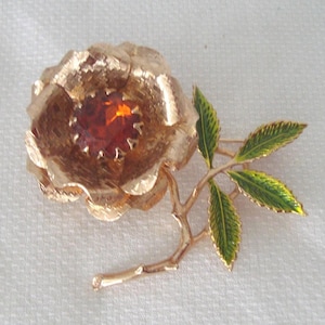 Può includere: Spilla a forma di rosa color oro con una pietra centrale color ambra. La spilla presenta petali dettagliati e uno stelo con foglie smaltate verdi e gialle. Un classico accessorio floreale.