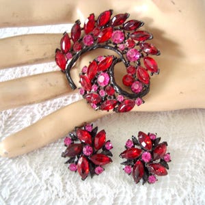 May include: A vintage jewelry set featuring a brooch and matching earrings. The brooch is a swirling design with red and pink gemstones set in a dark metal. The earrings are round, with a similar gemstone arrangement. The set is displayed on a lace surface.