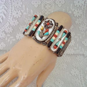 May include: A silver cuff bracelet featuring a central oval inlay of a flower design with mother-of-pearl, turquoise, coral, and jet stones. The bracelet includes side panels with geometric inlays of turquoise, coral, black, and white stone strips.