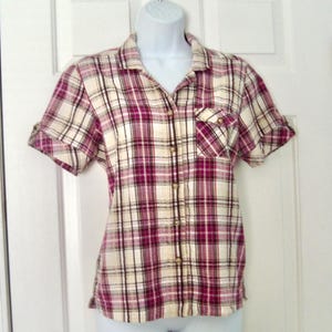 May include: A short-sleeved plaid shirt in cream, burgundy, and black. The shirt has a button-down front, a chest pocket, and rolled-up sleeves. The shirt is displayed on a white mannequin.