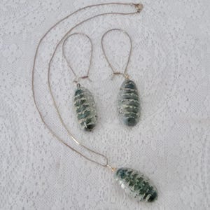 May include: A jewelry set consisting of a thin metal chain necklace and two matching drop earrings. Each piece features a smooth, oval-shaped pendant with a dark green and white banded pattern, suspended from simple silver-toned metal wire hooks.