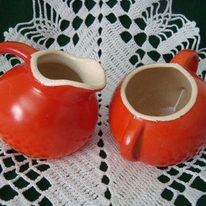 May include: Two bright orange ceramic jugs with rounded bodies and curved handles. One jug features a prominent pouring spout, while the other has a simple circular opening. Both vessels have a smooth, glossy finish and a light beige interior.