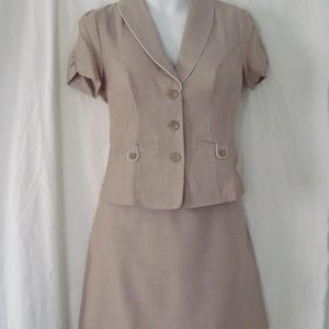 Puede incluir: Traje de dos piezas color beige. La chaqueta de manga corta tiene cuello con solapas con ribete blanco, tres botones y dos bolsillos delanteros. La falda a juego llega a la rodilla. El traje se exhibe en un maniquí.