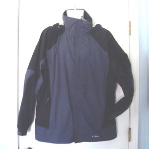 May include: A two-tone jacket in navy blue and black, with a high collar and a hood. The jacket has a zipper closure and the brand name "L.L.Bean" is visible on the lower right side. The jacket is hanging on a white hanger.