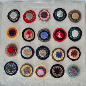 May include: A collection of twenty circular fabric patches arranged in a four-by-five grid. Each patch features layered, multi-colored textile circles with visible hand-stitched details in various thread colors, showcasing diverse textures and patterns.