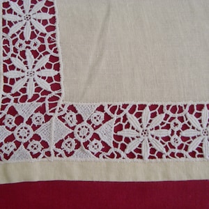 Może przedstawiać: Kremowa lniana tkanina z białym koronkowym obramowaniem. Koronka ma kwiatowy wzór z ażurowymi detalami. Tkanina jest prostokątna, z koronkowym wykończeniem wzdłuż krawędzi. Tło jest w kolorze głębokiej czerwieni.
