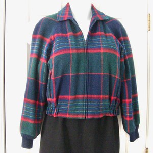 May include: A zip-up jacket featuring a plaid pattern in shades of dark green, navy blue, and bright red. The jacket has a collared neckline, long sleeves with ribbed cuffs, and a gathered elastic hemline at the waist, worn over a black garment.