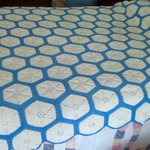 May include: A handmade crochet bedspread featuring a repeating pattern of white hexagonal motifs joined together by a vibrant blue yarn border. The intricate needlework creates a geometric design that covers the entire surface of the textile.
