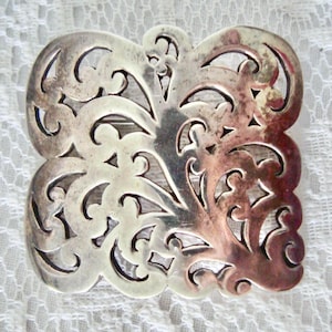 May include: A silver-toned metal brooch with an openwork design. The square brooch features a symmetrical pattern of swirling lines and cut-out shapes. The metal has a slightly tarnished appearance, with some areas showing a reddish hue.
