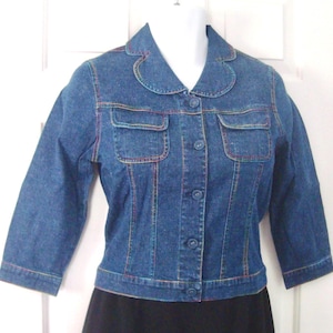 May include: A blue denim jacket with three-quarter sleeves, two front pockets, and a rounded collar. The jacket features colorful stitching along the seams and around the pockets. It has a button-up front and is displayed on a mannequin.