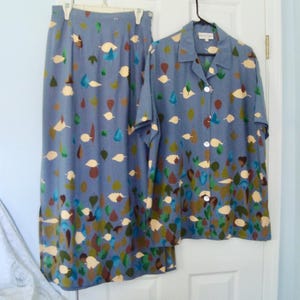 Pode incluir: Um conjunto de duas peças composto por uma camisa de manga curta com botões e uma saia comprida, ambas em tecido azul acinzentado com um padrão disperso de folhas creme, castanhas, verdes e azul-petróleo que se torna mais denso na bainha.