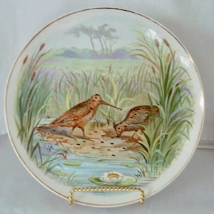 May include: A round ceramic plate with a gold-trimmed edge featuring a colorful illustration of two brown birds foraging in a marshy landscape with tall green reeds, cattails, and a single white water lily floating in the blue water below.
