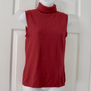 Può includere: Un top smanicato in maglia rossa con collo alto a lupetto. Il capo è esposto su un busto di manichino bianco, mostrando un colore rosso solido e una silhouette semplice e aderente con giromanica rifiniti e orlo dritto.