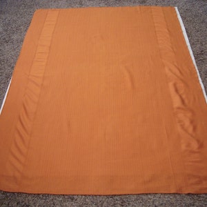 May include: A rectangular orange fabric panel featuring a subtle vertical pinstripe pattern throughout the center section. The fabric has a solid orange border on the left and right sides, with a thin white edge visible along the right side.