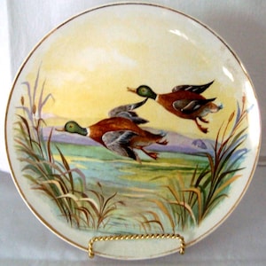 May include: A round ceramic decorative plate featuring a painted scene of two mallard ducks in flight over a marshy landscape with tall reeds, a calm body of water, and a soft yellow sky, finished with a thin gold rim around the circular edge.