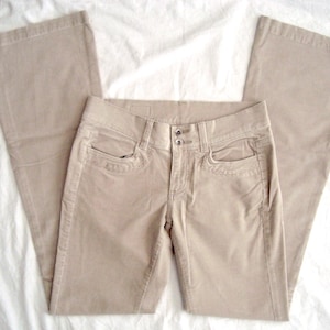 Può includere: Un paio di pantaloni in velluto a coste beige con gamba a zampa, vita bassa, due tasche anteriori e chiusura con doppio bottone centrale. I pantaloni presentano una vita strutturata con passanti per cintura e cuciture visibili lungo le gambe.