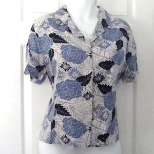 Puede incluir: Una camisa de manga corta con botones y estampado floral. La camisa presenta grandes rosas azules, hojas negras y flores más pequeñas blancas y grises sobre un fondo claro. El cuello y las mangas son visibles.
