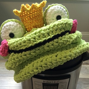 Può includere: Una rana di uncinetto verde con una corona gialla si trova sopra una pentola a pressione elettrica Instant Pot Plus argentata. Il display dell'Instant Pot mostra "8.8.8.8" e ha pulsanti per "Soffriggere/Grigliare", "Manuale", "Cottura a pressione", "Cottura lenta", "Yogurt", "Mantenere caldo", "Riso", "Multigrano", "Porridge", "Zuppa", "Stoufato", "Carne/Pollame", "Fagioli/Chili", "Torta", "Uovo", "Sterilizzare", "Caldo", "Manuale", "Regola", "Ritardo", "Più", "Meno", "Annulla", "Avvia", "Accensione/Spegnimento".