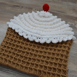 May include: A crocheted cupcake hat with a brown waffle stitch base and a white crocheted top with a red cherry on top.
