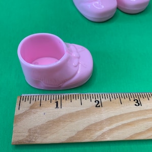 May include: Three pink plastic baby shoes with a bow detail. The shoes are about 1 inch long and are sitting on a green surface next to a wooden ruler.