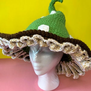 May include: A whimsical, handmade crochet hat in the shape of a mushroom. The hat features a green cap with white polka dots, a brown brim, and a ruffled edge in cream and brown. The hat is displayed on a white mannequin head against a yellow and pink background.