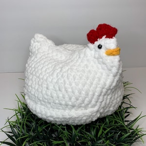 May include: White crocheted chicken with a red comb and yellow beak. The chicken is sitting on a bed of green grass.