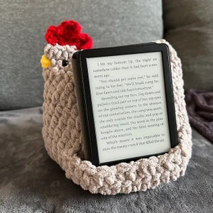 May include: A crocheted chicken-shaped book holder in beige and red holding a black e-reader. The e-reader screen displays text from a fantasy novel.