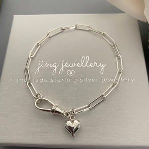 May include: A silver paperclip chain bracelet with a heart charm. The bracelet features a lobster clasp and is presented on a light grey box. The text "jing jewellery" and "Handmade sterling silver jewellery" are visible.