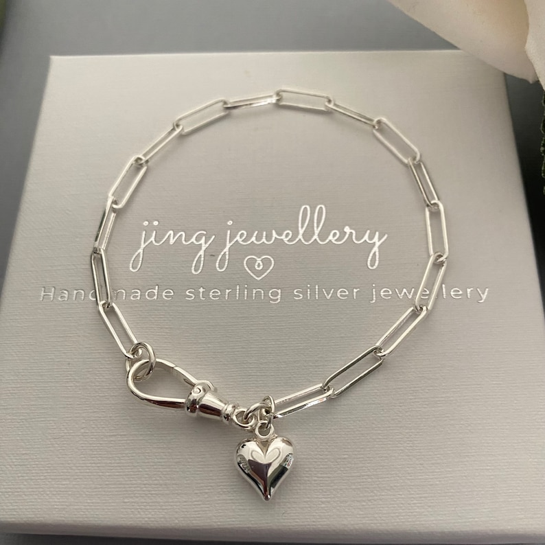 May include: A silver paperclip chain bracelet with a heart charm. The bracelet features a lobster clasp and is presented on a light grey box. The text "jing jewellery" and "Handmade sterling silver jewellery" are visible.