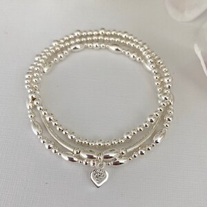May include: A set of three silver bracelets with a small heart charm. The bracelets are made of small silver beads and are layered on top of each other.