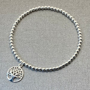 May include: A silver beaded bracelet featuring a tree of life charm. The bracelet is made from small, round beads and has a delicate, feminine design.
