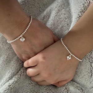 May include: Two silver beaded bracelets with heart charms. The bracelets are on the wrists of two people, one adult and one child.