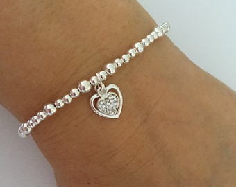 Puede incluir: Una pulsera de plata con un dije en forma de corazón. El dije en forma de corazón está lleno de pequeños cristales transparentes.