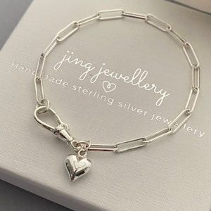 May include: A silver paperclip chain bracelet with a heart charm. The bracelet has a lobster clasp and a small heart pendant with a heart-shaped cutout. The packaging displays the text "jing jewellery".