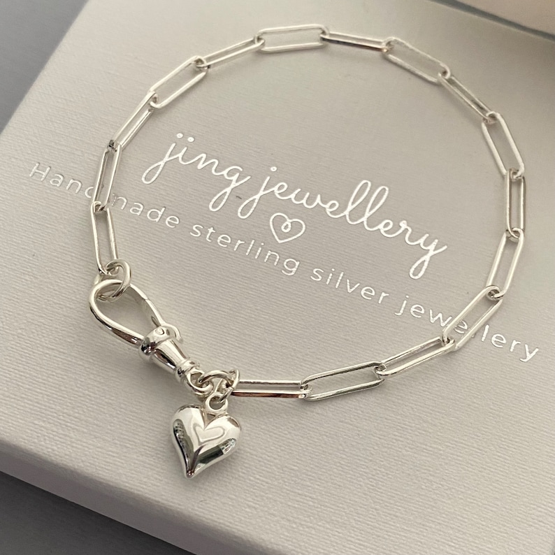 May include: A silver paperclip chain bracelet with a heart charm. The bracelet has a lobster clasp and a small heart pendant with a heart-shaped cutout. The packaging displays the text "jing jewellery".