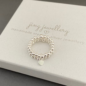 May include: A silver ring with a heart charm. The ring is made of small beads and is stacked on top of itself. The ring is in a white box with the text "jing jewellery handmade sterling silver jewellery".
