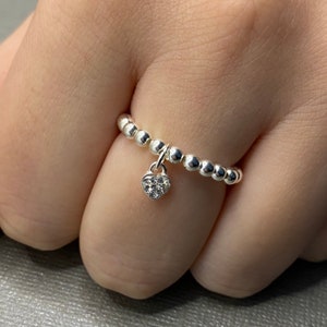 May include: A silver ring with a small, clear heart charm dangling from it. The ring is made of small, round beads.