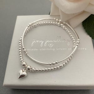 May include: Two silver bracelets with a heart charm. The bracelets are made of small beads and are stacked on top of each other. The bracelets are in a white box with the text "jing jewellery" and "handmade sterling silver jewellery".