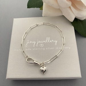May include: A silver link bracelet with a heart pendant. The bracelet is presented on a white box with the text "jing jewellery" and "Handmade sterling silver jewellery". A white rose is in the background.