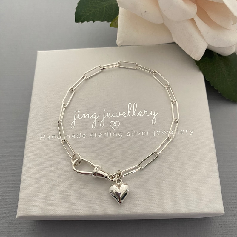 May include: A silver link bracelet with a heart pendant. The bracelet is presented on a white box with the text "jing jewellery" and "Handmade sterling silver jewellery". A white rose is in the background.
