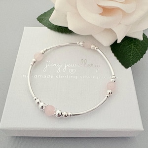 May include: A silver bangle bracelet featuring pink gemstone beads. The bracelet rests on a white box with the text "jing jewellery" and "Handmade sterling silver jewellery". A white rose is in the background.