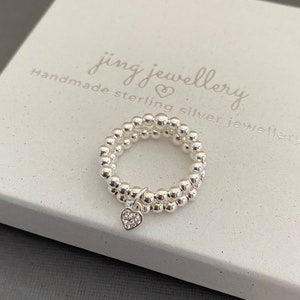 May include: A silver ring with a heart charm. The ring is made of small beads and the charm has a small crystal in the centre. The ring is on a white box with the text "jing jewellery handmade sterling silver jewellery".