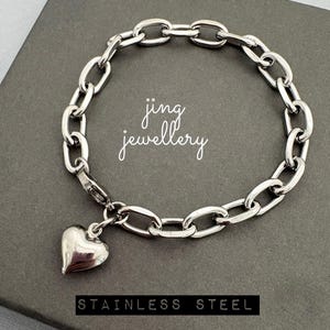 May include: A silver stainless steel bracelet with a heart charm. The bracelet has a chain link design and the text "jing jewellery" is written in cursive. The words "STAINLESS STEEL" are written in a black rectangle.