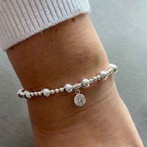 May include: A silver beaded bracelet with a clear crystal charm. The bracelet is on a person's wrist.