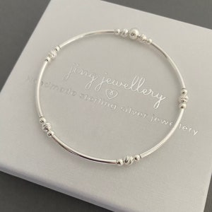 May include: A silver bangle bracelet with small beads and a larger bead in the centre. The bracelet is on a white background with the text "Jing Jewellery Handmade Sterling Silver Jewellery".