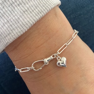 May include: A silver bracelet with a heart-shaped charm. The bracelet has a chain link design and a clasp. The heart charm is polished silver. The bracelet is worn on a wrist.